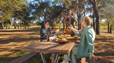 Bir grup arkadaş güneşli bir ormanda pikniğin tadını çıkarıyorlar. Önlerinde yiyecek ve içeceklerle ahşap bir masada oturuyorlar, neşeli ve rahat bir açık hava yaratıyorlar..