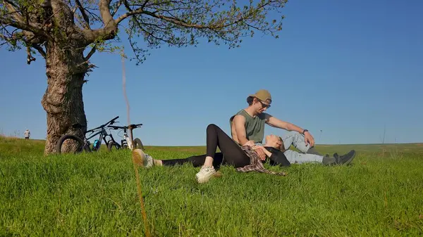 Bir çift, yakınlarda bisikletleri olan sakin yeşil bir tarlada bir ağacın altında oturuyor. Güneşli bir gün huzurlu bir sahne yaratır..