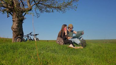 Bir çift bir ağacın altında oturur, uçsuz bucaksız yeşil bir alanda bisiklet maceraları sırasında haritayı keşfederler..