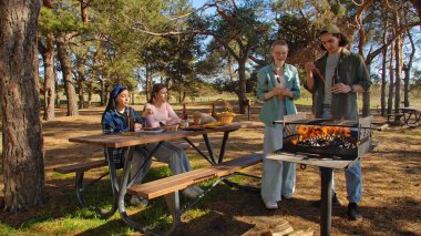 Bir grup arkadaş güneşli bir günün tadını çıkarıyorlar. Uzun ağaçlarla çevrili, pitoresk bir parkta barbekü yapıyorlar..