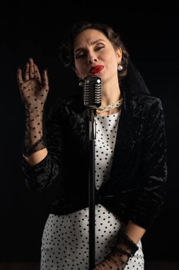 Geçmişe dönük giyinmiş bir kadın, eskimiş bir mikrofona koyu bir arka planda tutkuyla şarkı söylüyor..