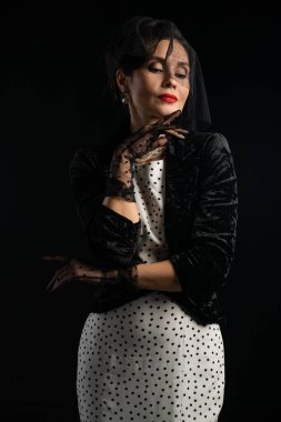 Eskimiş siyah kadife ceket ve beyaz benekli elbise giymiş sofistike bir kadın, bir peçe ve inci, koyu bir zemin karşı set.