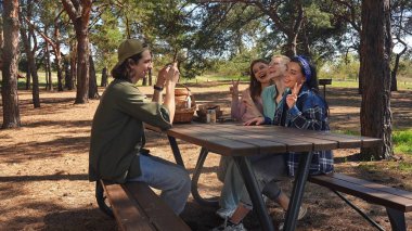 Bir grup arkadaş ormanda pikniğin tadını çıkarıp anı fotoğrafla yakalıyorlar. Doğayla iç içe olup, bir piknik sepetiyle ahşap bir masanın etrafında toplanırlar..