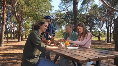 Bir grup arkadaş güneşli bir ormanda piknik yapıyorlar, sohbet ediyorlar ve açık havada taze sebzelerin tadını çıkarıyorlar..