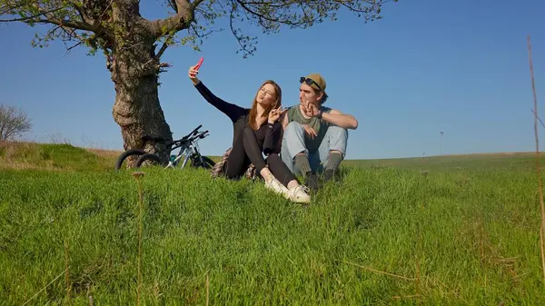 Genç bir çift yeşil bir tarlada bir ağacın altında otururken selfie çekiyor, açık gökyüzünün altında bisiklet sürüyor..