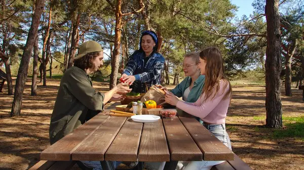 Bir grup arkadaş güneşli bir ormanda piknik yapıyorlar, sohbet ediyorlar ve açık havada taze sebzelerin tadını çıkarıyorlar..