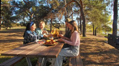 Bir grup arkadaş güneşli bir ormanda pikniğin tadını çıkarıyorlar. Önlerinde yiyecek ve içeceklerle ahşap bir masada oturuyorlar, neşeli ve rahat bir açık hava yaratıyorlar..