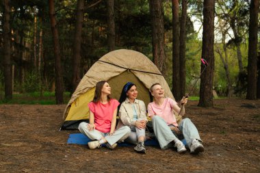 Ormandaki bir çadırın önünde oturan üç arkadaş selfie çubuğuyla anılarını yakalıyor, dışarıda eğleniyorlar..
