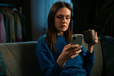 Bir kadın rahat bir şekilde kanepede oturur, akıllı telefonuyla uğraşırken kahvesini yudumlar, rahatlamanın tadını çıkarır..