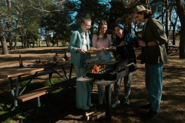 Bir grup arkadaş güneşli bir günün tadını çıkarıyorlar. Uzun ağaçlarla çevrili, pitoresk bir parkta barbekü yapıyorlar..