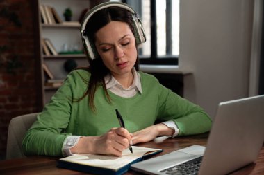 Odaklanmış bir kadın laptopuyla çalışır, kulaklık takar ve modern ofis ortamında notlar alır..