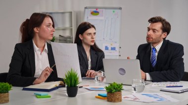 Modern bir ofiste yapılan toplantı sırasında resmi giyinmiş üç profesyonelden oluşan bir grup iş raporlarını inceliyor..