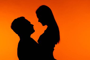 Romantik poz veren bir çiftin silüeti canlı turuncu bir arkaplanda, samimiyet ve bağlantıyı vurguluyor..