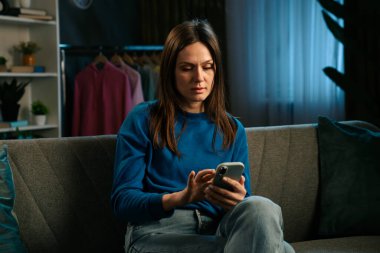 Kanepede oturan ve rahat bir oturma odasında akıllı telefonuyla video izleyen bir kadın..