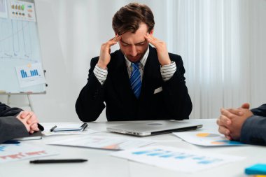 Takım elbiseli bir işadamı, evrak ve çizelgelerle çevrili bir ofis toplantısında stresli görünüyor..