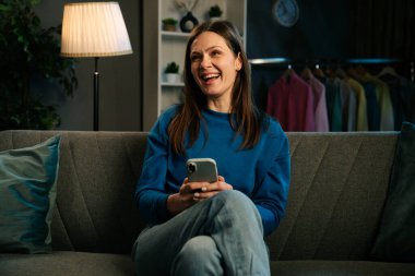 Neşeli bir kadın kanepede oturur ve rahat bir oturma odasında akıllı telefonuyla video izler..