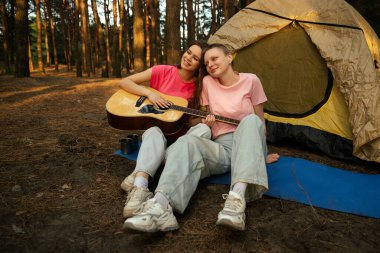 İki arkadaş ormanda bir çadırın yanında dinleniyor gitar çalıyor ve açık havada huzurlu bir ortam seyrediyor..