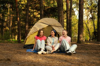 Üç kişi bir orman ortamında çadırın önünde mutlu bir şekilde oturur ve birlikte ferahlatıcı bir kamp deneyiminin tadını çıkarırlar..
