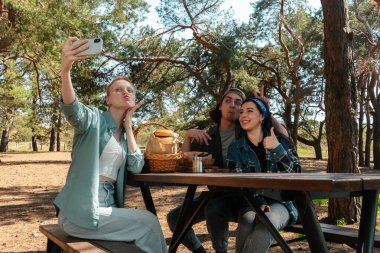 Neşeli bir arkadaş grubu parkta pikniğin tadını çıkarıyor. Uzun ağaçların altında selfie çekiyorlar..