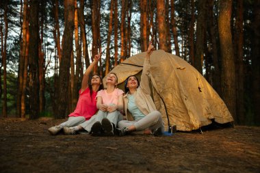Yoğun bir ormanda çadırda kamp maceralarının keyfini çıkarırken üç kadın gökyüzünü neşeyle işaret ediyor..