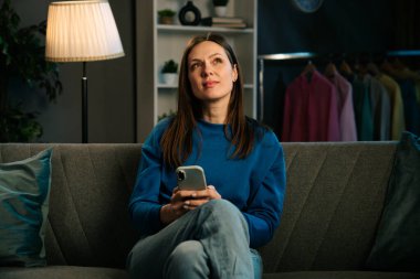 Kanepede oturan ve rahat bir oturma odasında akıllı telefonuyla video izleyen bir kadın..