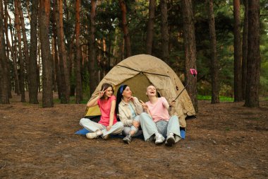 Ormandaki bir çadırın önünde oturan üç arkadaş selfie çubuğuyla anılarını yakalıyor, dışarıda eğleniyorlar..
