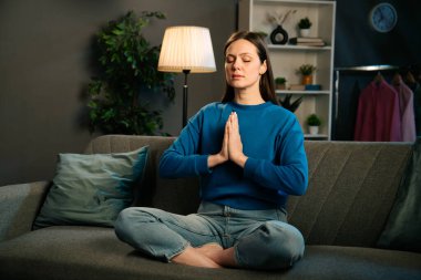 Huzurlu bir oturma odasındaki kanepede meditasyon yapan bir kadın. Etrafı bitkilerle çevrili ve dinlenmek için hafif ışıklandırmalı..