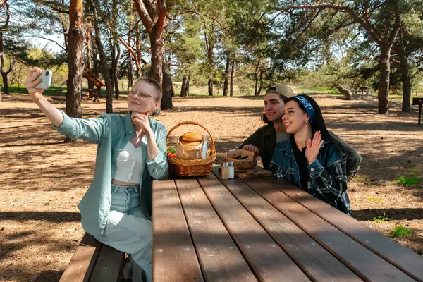 Bir grup arkadaş ormanda pikniğin tadını çıkarıyor ve anı selfie ile yakalıyorlar. Doğayla iç içe olup, bir piknik sepetiyle ahşap bir masanın etrafında toplanırlar..