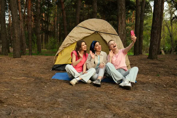 Üç arkadaş ormanda bir çadırın önünde oturmuş, gülüyor ve selfie çekiyor, açık hava macerasının zevkini yakalıyorlar..