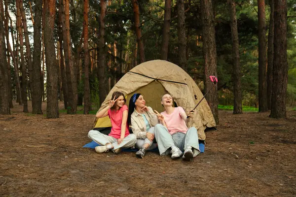 Ormandaki bir çadırın önünde oturan üç arkadaş selfie çubuğuyla anılarını yakalıyor, dışarıda eğleniyorlar..