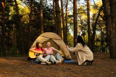 Arkadaşlarla dolu canlı bir kamp sahnesi müzik ve kahkahaların tadını çıkaran yemyeşil bir ormanda doğa ile çevrili..
