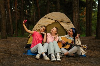 Üç arkadaş ormanda kamp yapmaktan zevk alıyor. Çadırlarının dışında selfie çekiyorlar ve gitar çalıyorlar..