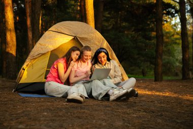 Üç arkadaş ormanda bir çadırın yanında oturmuş, bir dizüstü bilgisayarın üzerinde yürüyüş ve görüntülü sohbetin tadını çıkarıyorlar..