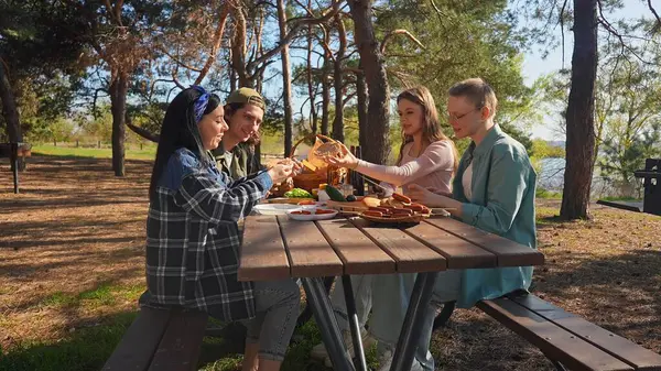 Bir grup arkadaş güneşli bir ormanda pikniğin tadını çıkarıyorlar. Önlerinde yiyecek ve içeceklerle ahşap bir masada oturuyorlar, neşeli ve rahat bir açık hava yaratıyorlar..