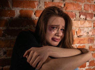 Yaralı genç bir kadın, kasvetli bir ortamda üzüntüsünü ve üzüntüsünü ifade eden tuğla bir duvara yaslanır..