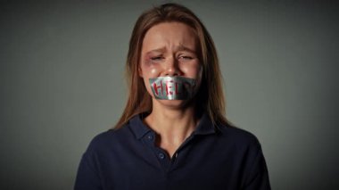 Yüzü morarmış bir kadın ağzını bantla kapatarak ağlıyor ve çaresizce yardıma ihtiyacı olduğunu söylüyor. Yavaş çekim.
