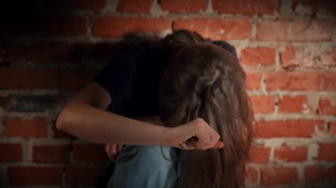 Yaralı genç bir kadın, kasvetli bir ortamda üzüntüsünü ve üzüntüsünü ifade eden bir tuğla duvarın karşısında oturuyor. Yavaş çekim.