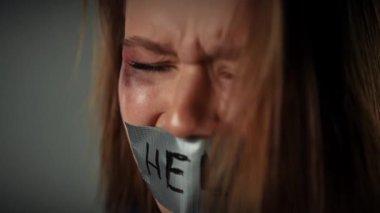 Yüzü morarmış bir kadın ağzını bantla kapatarak ağlıyor ve çaresizce yardıma ihtiyacı olduğunu söylüyor. Yavaş çekim.