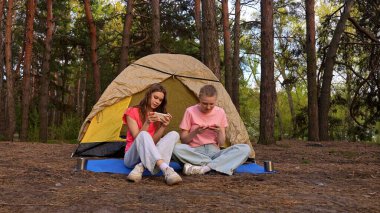 İki arkadaş çadırın yanında oturur, sohbet eder ve akıllı telefonlarıyla oyun oynarlar, ormanda yürüyüş yaparlar..
