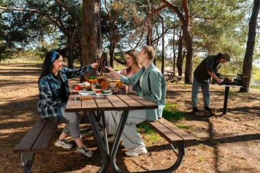 Orman ortamında piknik yapan bir grup arkadaş, ahşap bir masada kahkaha ve yemek paylaşıyorlar..