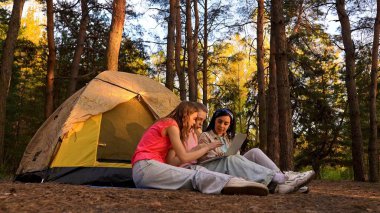 Üç arkadaş ormanda bir çadırın yanında oturmuş, bir dizüstü bilgisayarın üzerinde yürüyüş ve görüntülü sohbetin tadını çıkarıyorlar..
