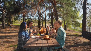 Bir grup arkadaş güneşli bir ormanda pikniğin tadını çıkarıyorlar. Önlerinde yiyecek ve içeceklerle ahşap bir masada oturuyorlar, neşeli ve rahat bir açık hava yaratıyorlar..