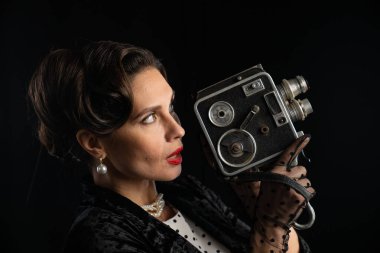 Retro giysili bir kadın klasik bir kamera tutar, klasik bir cazibe ve zarafet yayar, karanlık bir arka plana karşı..