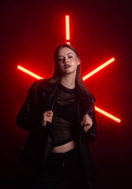 Arka planda kırmızı ışıklar yanıp sönen siyah deri ceketli bir kadın poz veriyor. Atmosfer gergin ve modern. Moda yazıları, giyim markaları veya aksesuar reklamları için..