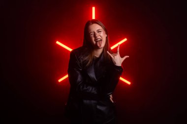 Deri ceketli genç bir kadın cesur, neon ışıklı bir arka plana karşı neşeli bir ifadeyle poz veriyor. Moda yazıları, giyim markaları veya aksesuar reklamları için..