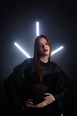 Bir kadın karanlık bir ortamda, parlak neon ışıklarla aydınlanarak güvenle ayakta duruyor. Onun cesur duruşu ve katıksız giyimi modern estetiği güçlendirir. Moda yazıları, giyim markaları veya aksesuar reklamları için..