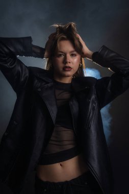 Bir kadın siyah deri ceketle rahatça poz veriyor. Dramatik ışıklandırma gizemli bir atmosfer yaratır. Moda yazıları, giyim markaları veya aksesuar reklamları için..