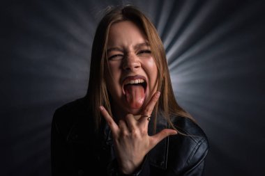 Siyah deri ceketli bir kadın dilini uzatır ve el hareketi yapar. Karanlık bir zemine karşı enerji ve heyecan taşır. Moda yazıları, giyim markaları veya aksesuar reklamları için..