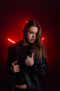 Siyah deri ceketli genç bir kadın dramatik bir kırmızı ışık arka planına karşı güvenle poz veriyor. Moda yazıları, giyim markaları veya aksesuar reklamları için..