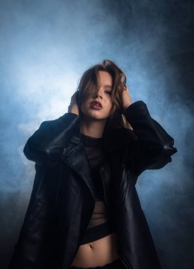 Siyah deri ceketli genç bir kadın, dumanlı mavi bir arka plana karşı özgüvenle poz veriyor, çok tarz ve tavır sergiliyor. Moda yazıları, giyim markaları veya aksesuar reklamları için..